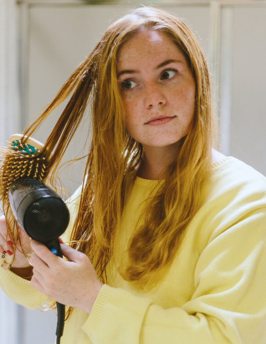 Teenage girl with red hair drying her wet hair with a hairdryer and brush.