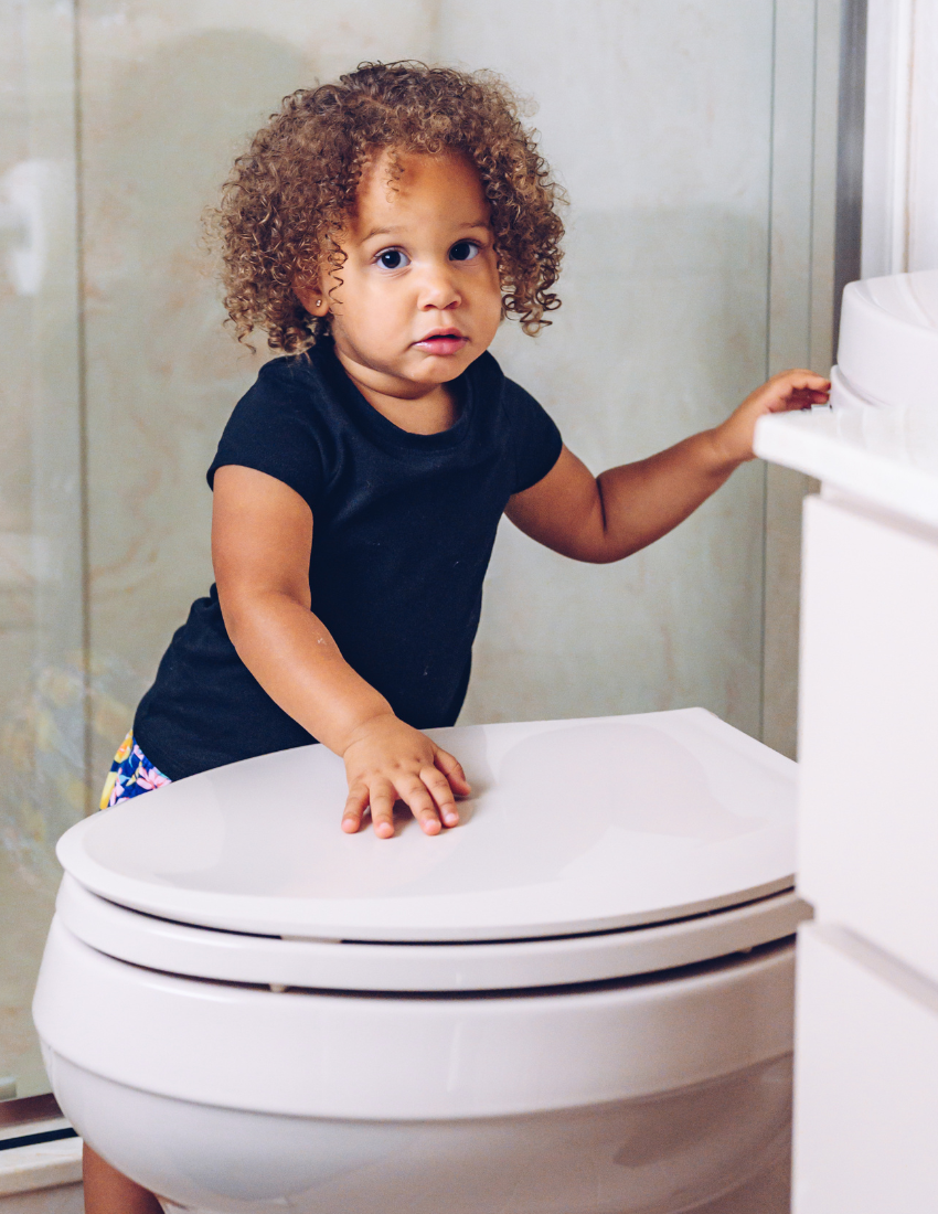 A toddler with curly hair, dressed in a black shirt, stands in the bathroom with one hand resting on a closed white toilet lid and the other hand touching a nearby surface. In the background, there is a shower with a glass door. The child looks toward the camera with a neutral expression. This moment is part of their journey featured in "Toileting Masterclass" by The Play Way.