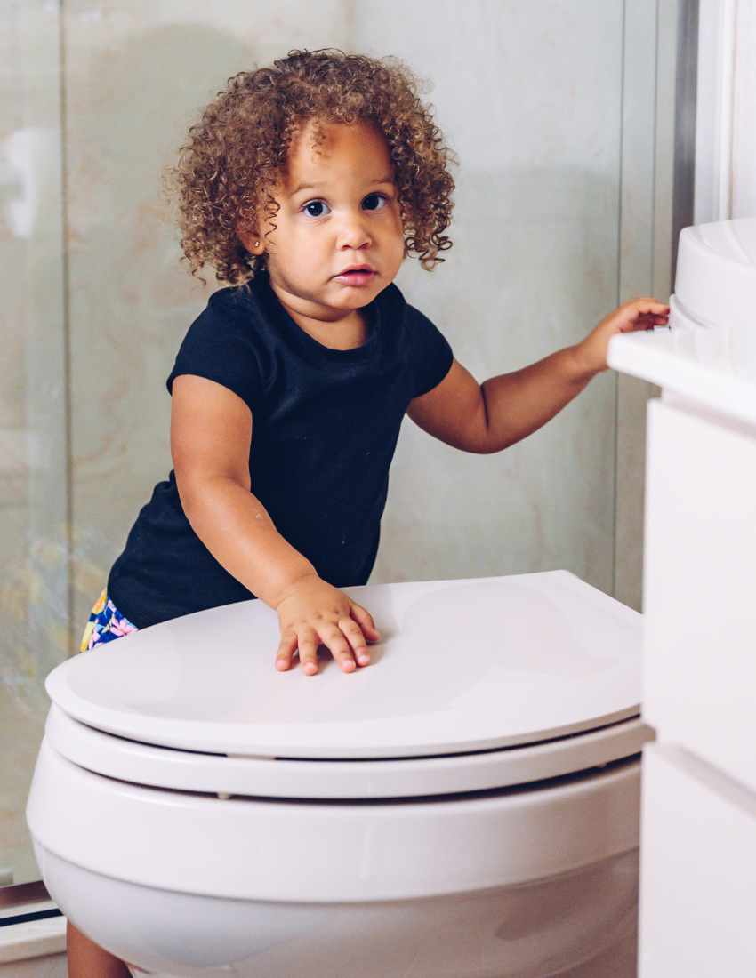 Young child with curly hair standing by a toilet.