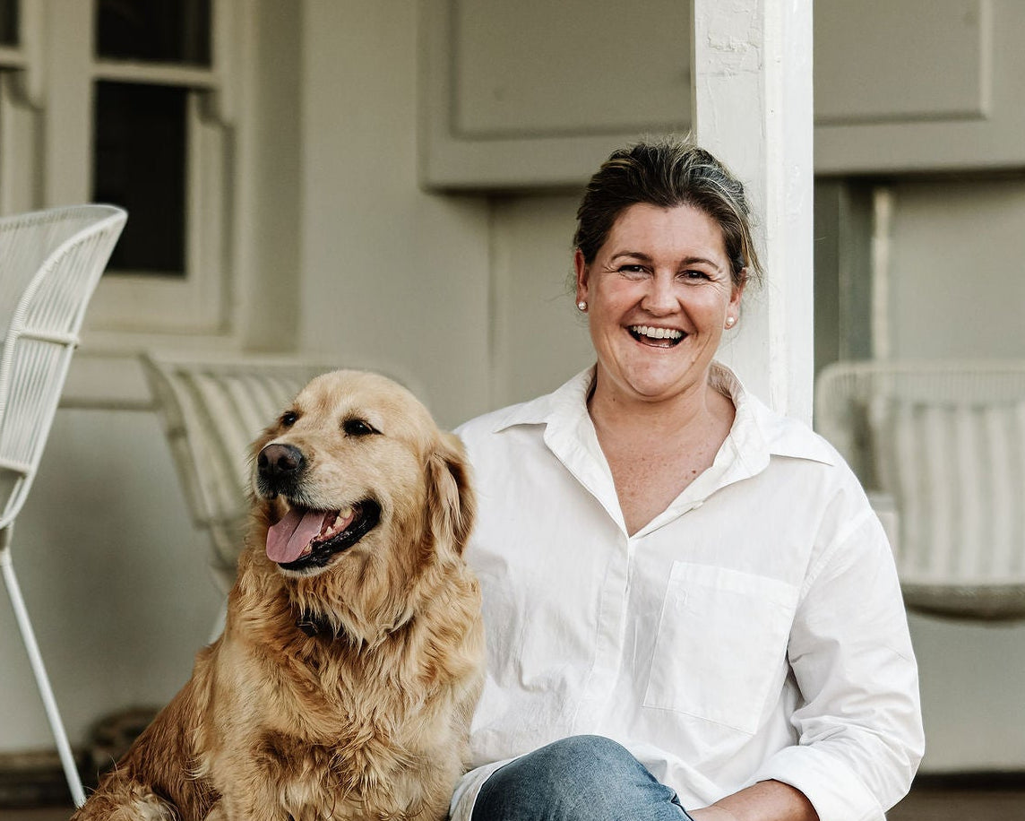 Kait in a white button-down shirt and blue jeans sits smiling on a porch step next to a golden retriever dog. The porch has white columns and light-colored cabinets in the background.