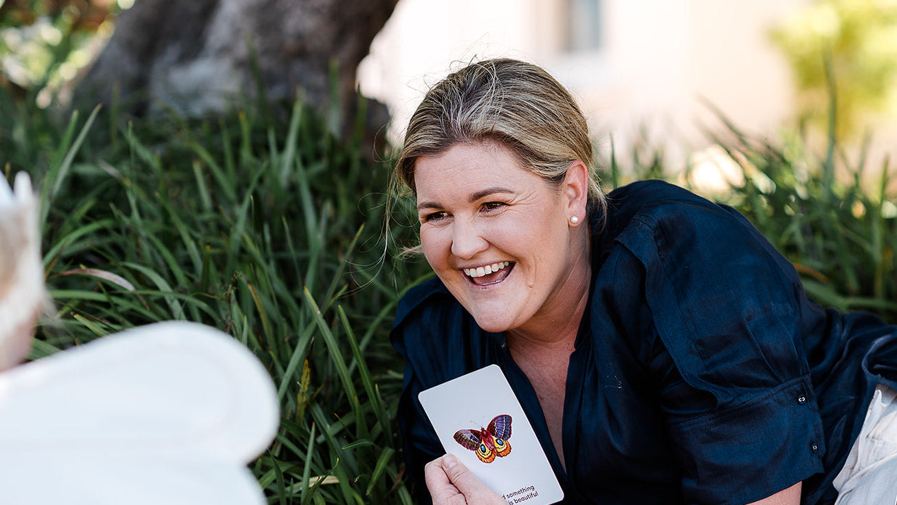 Kait Cummins lying on the ground holding a card with a butterfly design, surrounded by greenery.