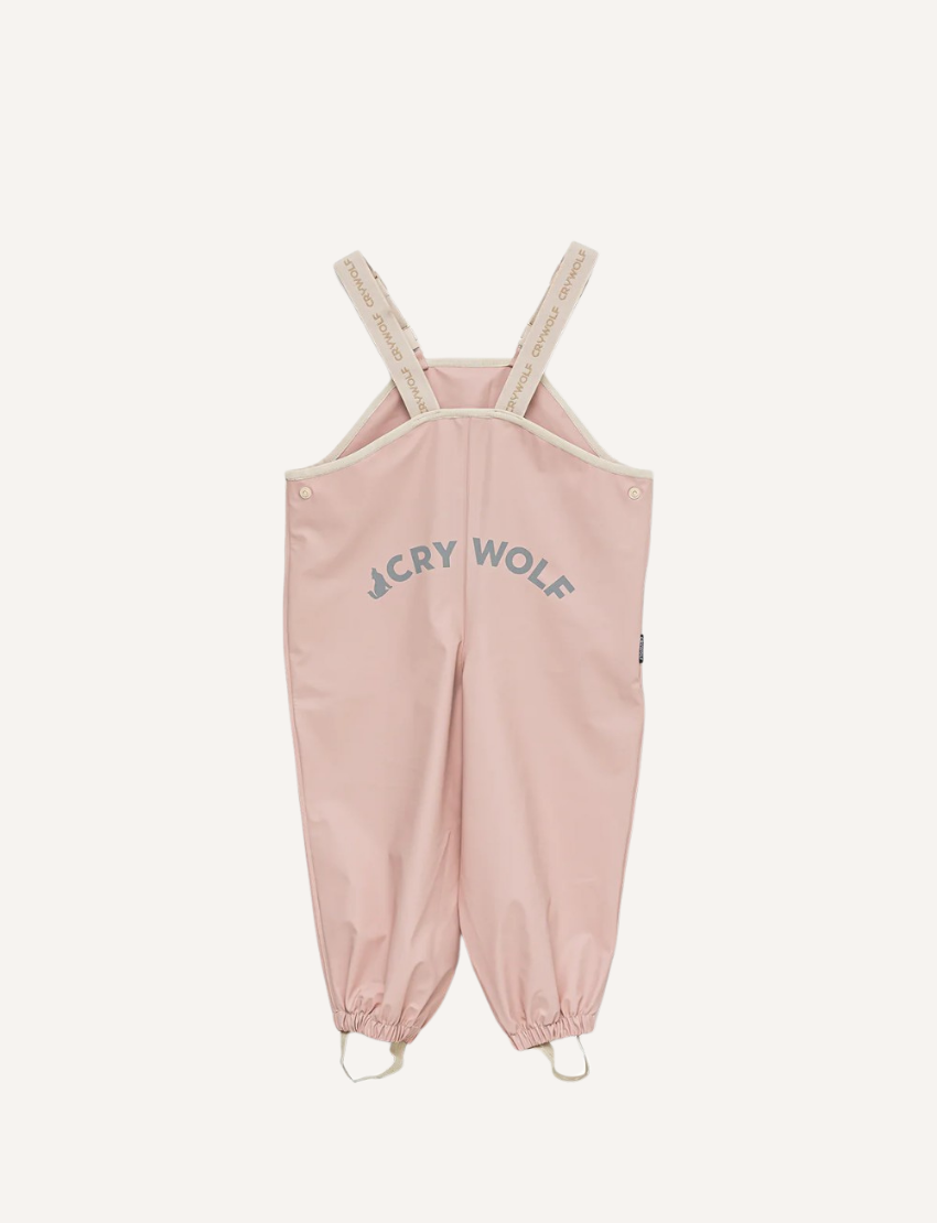 This image shows the back view of the Cry Wolf Rain Overalls in Dusty Pink, designed for children aged 3 years. The overalls feature elasticated cuffs at the ankles and adjustable shoulder straps with the brand's name printed on them. The words "A CRY WOLF" are printed across the back.