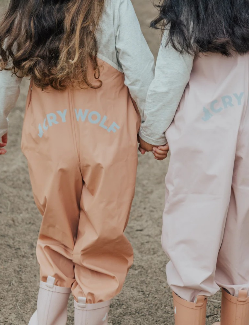 This image shows the back view of the Cry Wolf Rain Overalls in Dusty Pink, designed for children aged 3 years. The overalls feature elasticated cuffs at the ankles and adjustable shoulder straps with the brand's name printed on them. The words "A CRY WOLF" are printed across the back.