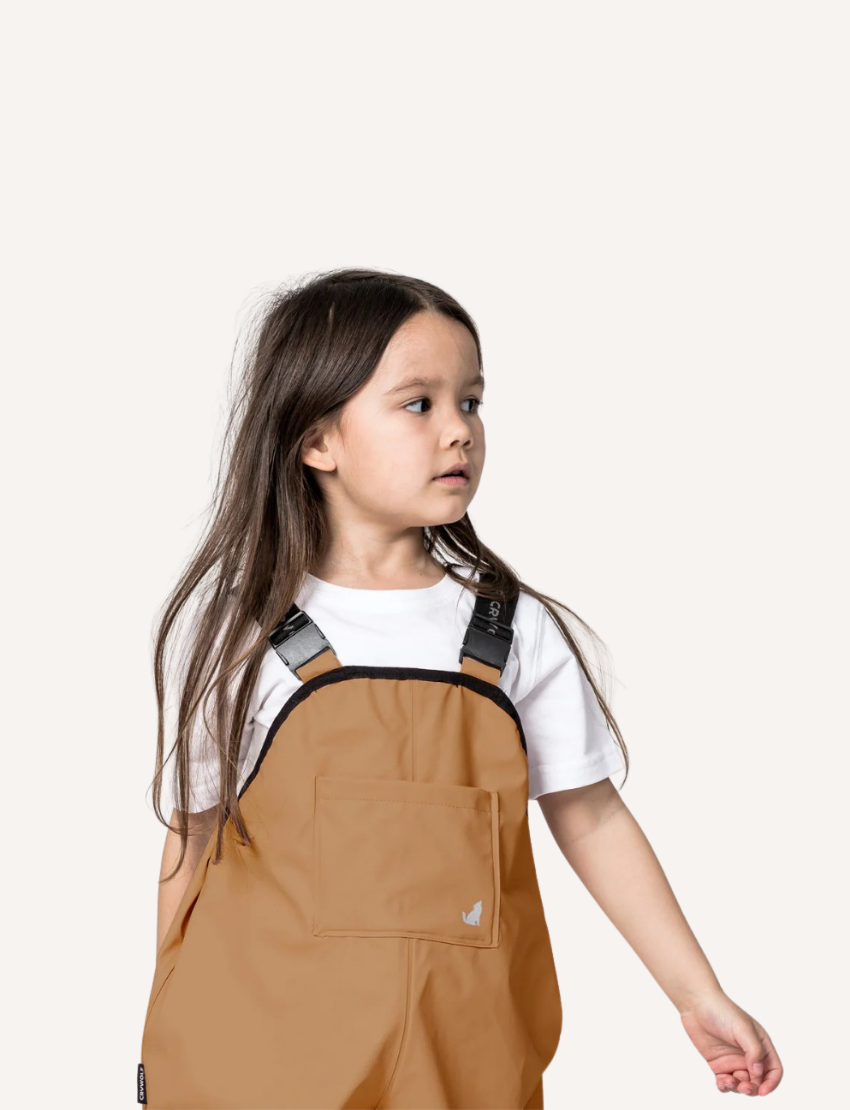 A pair of children's tan waterproof Rain Overalls from Cry Wolf with elasticated ankle cuffs, black adjustable shoulder straps, and the words "Cry Wolf" printed in gray on the back. There are black foot straps at the bottom of each leg opening.