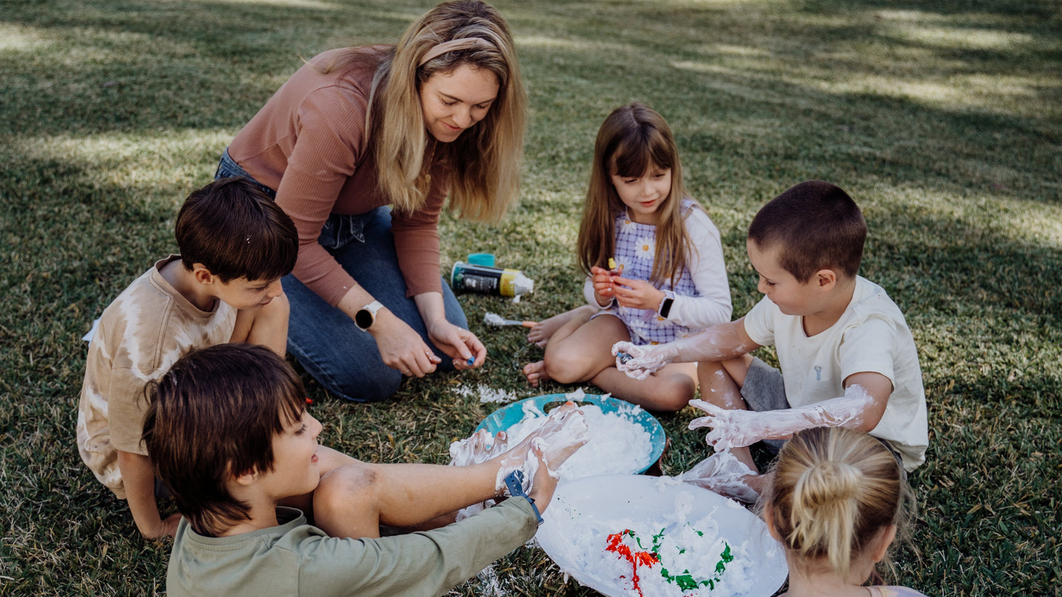 Group of children and a woman sitting on grass outdoors, engaged in an activity.