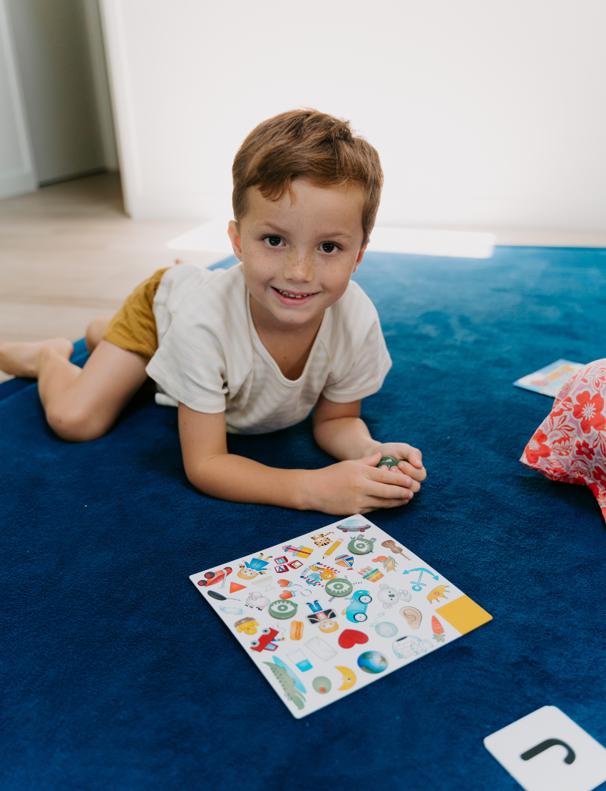 A colorful "Sound Awareness Program" box from The Play Way is open, revealing educational toys and books for phonological awareness. Inside are "CVC Word Builders," "Spot Markers," and "Slimy Rhymes," all in vibrant packaging designed to inspire early handwriting skills.