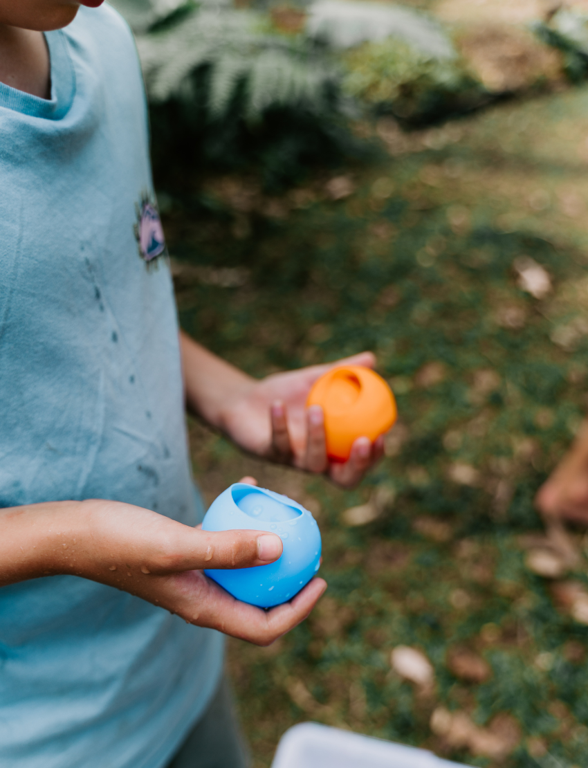A person in a light blue shirt holds a blue ball in their left hand and an orange ball in their right. The scene, featuring green grass and fallen leaves, embodies the essence of The Play Way's Summer Holidays Program.