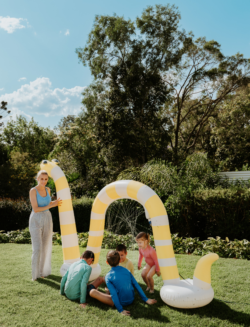 A person in a light blue shirt holds a blue ball in their left hand and an orange ball in their right. The scene, featuring green grass and fallen leaves, embodies the essence of The Play Way's Summer Holidays Program.