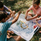 A person in a light blue shirt holds a blue ball in their left hand and an orange ball in their right. The scene, featuring green grass and fallen leaves, embodies the essence of The Play Way's Summer Holidays Program.
