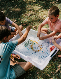 The Therapy At Home Summer Holidays Program comes in a green box with colorful abstract shapes and includes rackets, a camera, an ice cube tray for water play, craft supplies, cards, an air pump, and activity booklets—all neatly displayed upright.