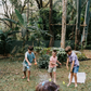 A person in a light blue shirt holds a blue ball in their left hand and an orange ball in their right. The scene, featuring green grass and fallen leaves, embodies the essence of The Play Way's Summer Holidays Program.