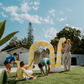 A person in a light blue shirt holds a blue ball in their left hand and an orange ball in their right. The scene, featuring green grass and fallen leaves, embodies the essence of The Play Way's Summer Holidays Program.
