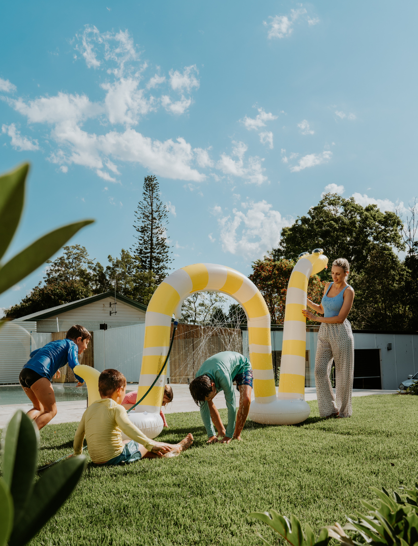 A person in a light blue shirt holds a blue ball in their left hand and an orange ball in their right. The scene, featuring green grass and fallen leaves, embodies the essence of The Play Way's Summer Holidays Program.