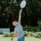 A person in a light blue shirt holds a blue ball in their left hand and an orange ball in their right. The scene, featuring green grass and fallen leaves, embodies the essence of The Play Way's Summer Holidays Program.