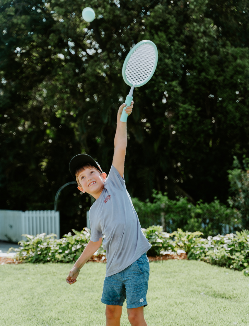 A person in a light blue shirt holds a blue ball in their left hand and an orange ball in their right. The scene, featuring green grass and fallen leaves, embodies the essence of The Play Way's Summer Holidays Program.
