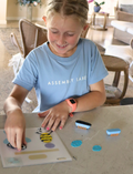 The Therapy At Home STEM: 3D Printing Pen Program includes a green pen, booklets, filaments, and kits to create running animals and walking robots—ideal for fine motor skills and STEM learning. The box is decorated with colorful abstract shapes.