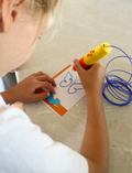 The Therapy At Home STEM: 3D Printing Pen Program includes a green pen, booklets, filaments, and kits to create running animals and walking robots—ideal for fine motor skills and STEM learning. The box is decorated with colorful abstract shapes.