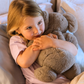 A parent with two kids is on a bed. They and one child are exploring The Play Way's Sleep Program box, while the other child holds a book. The bedroom features a decorative headboard and a patterned lamp.