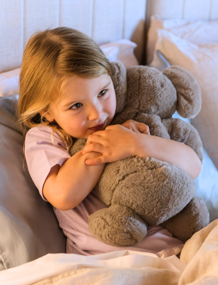 A parent with two kids is on a bed. They and one child are exploring The Play Way's Sleep Program box, while the other child holds a book. The bedroom features a decorative headboard and a patterned lamp.