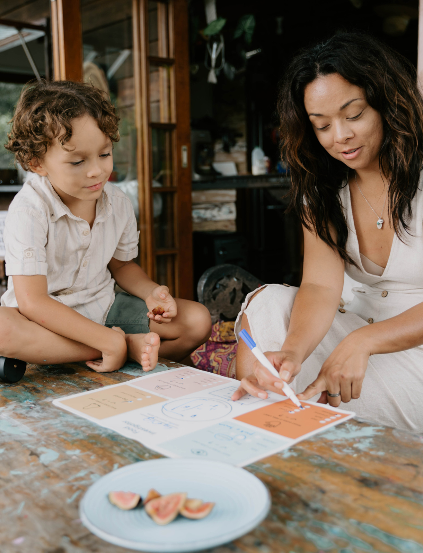 A child with curly hair and an adult next to them smile while pointing at a laptop displaying the Routines Program by The Play Way. The table is covered with small picture cards, and a guitar, couch, and painting are visible in the room.