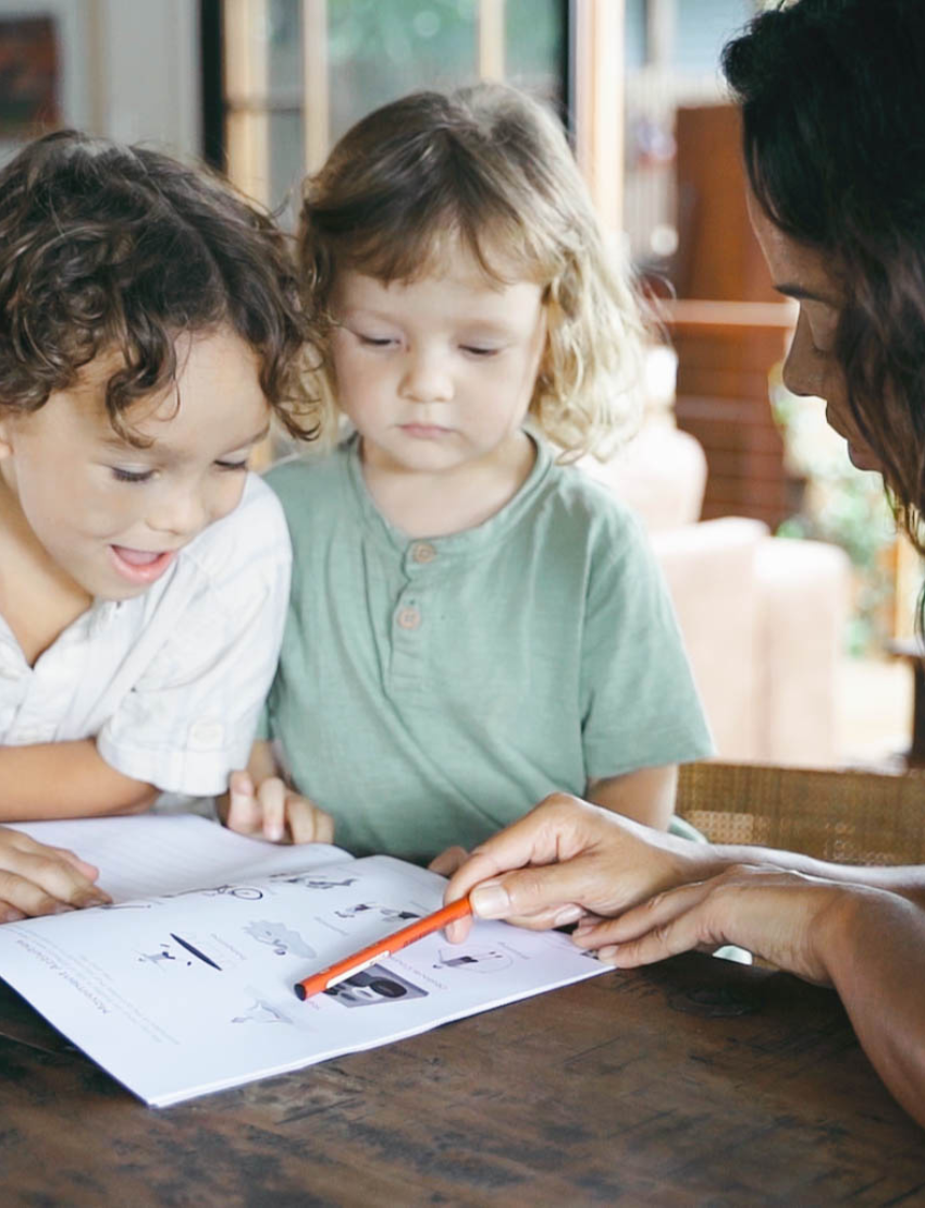 A child with curly hair and an adult next to them smile while pointing at a laptop displaying the Routines Program by The Play Way. The table is covered with small picture cards, and a guitar, couch, and painting are visible in the room.