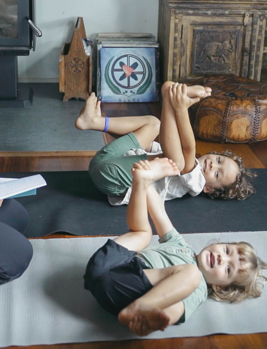 A child with curly hair and an adult next to them smile while pointing at a laptop displaying the Routines Program by The Play Way. The table is covered with small picture cards, and a guitar, couch, and painting are visible in the room.