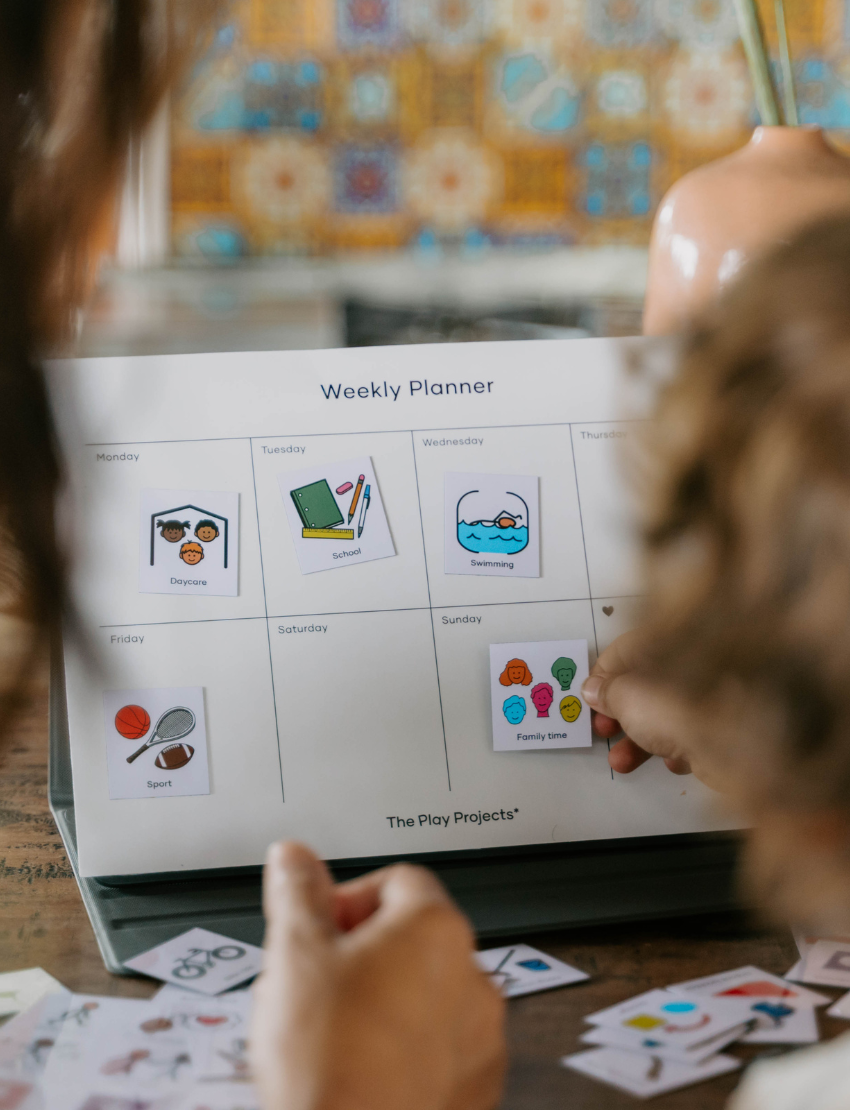 A child with curly hair and an adult next to them smile while pointing at a laptop displaying the Routines Program by The Play Way. The table is covered with small picture cards, and a guitar, couch, and painting are visible in the room.