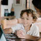 A child with curly hair and an adult next to them smile while pointing at a laptop displaying the Routines Program by The Play Way. The table is covered with small picture cards, and a guitar, couch, and painting are visible in the room.