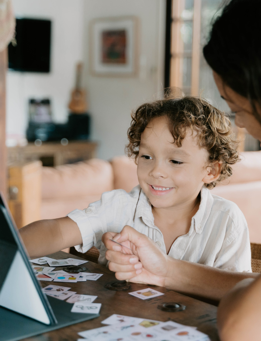 A child with curly hair and an adult next to them smile while pointing at a laptop displaying the Routines Program by The Play Way. The table is covered with small picture cards, and a guitar, couch, and painting are visible in the room.