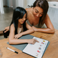 A woman and a girl sit at a wooden table, with the girl pointing at a dry erase board featuring The Play Way's Self-Care Program content. A marker lies nearby. They are in a modern home characterized by light-colored walls and furnishings.