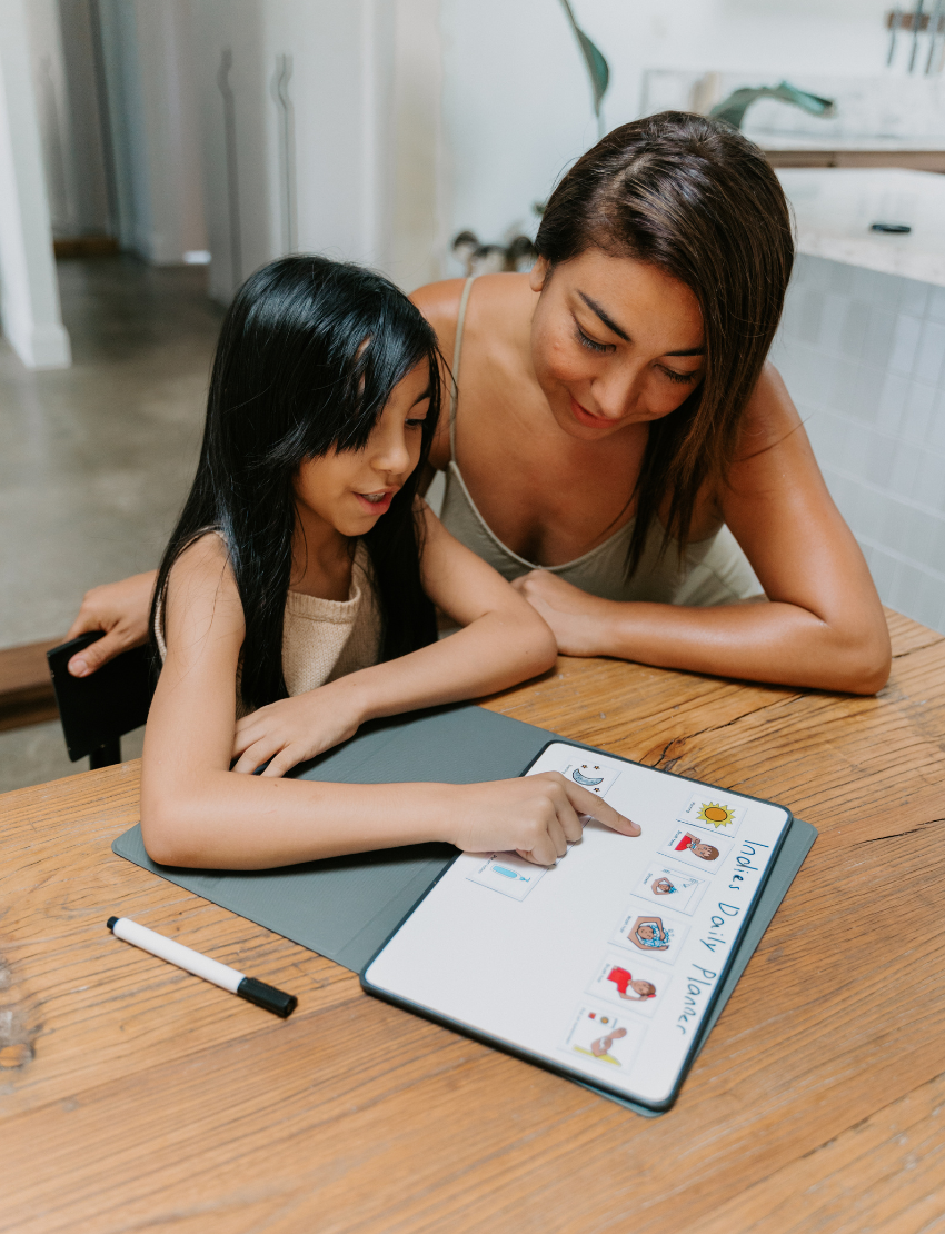 A woman and a girl sit at a wooden table, with the girl pointing at a dry erase board featuring The Play Way's Self-Care Program content. A marker lies nearby. They are in a modern home characterized by light-colored walls and furnishings.