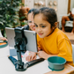 Two children sit at a table; one in a white shirt uses The Play Way's green tool to cut strawberries on a wooden board, focusing intently. The other wears yellow, watching with interest as strawberry pieces sit nearby. Part of the Feeding and Mealtimes: Helping You Get Started Program.