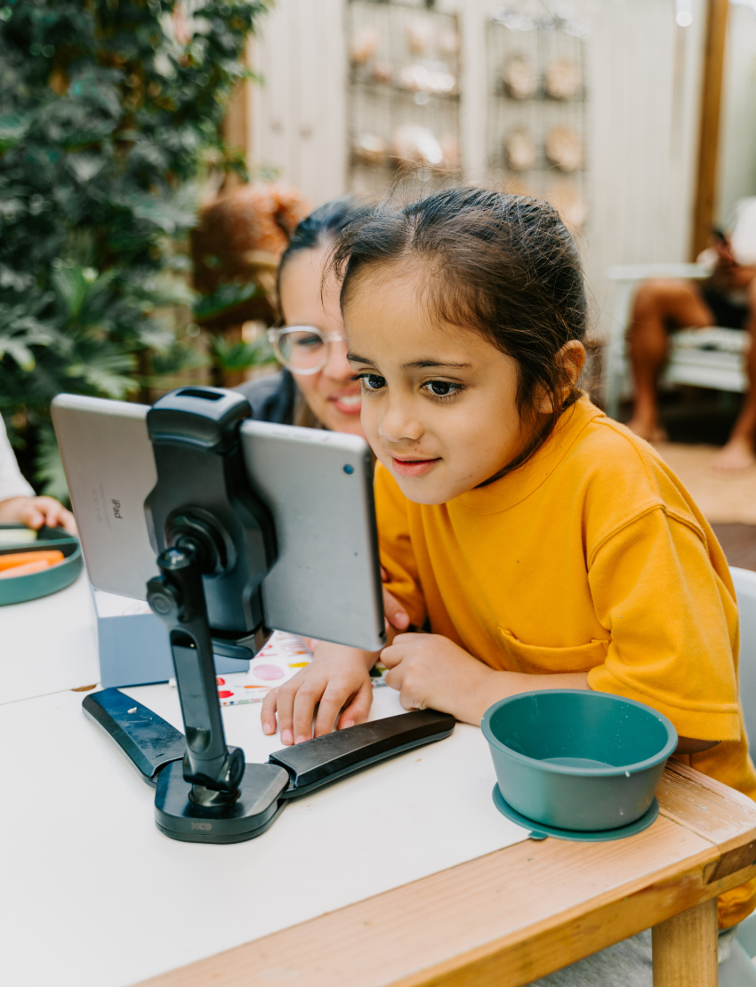 Two children sit at a table; one in a white shirt uses The Play Way's green tool to cut strawberries on a wooden board, focusing intently. The other wears yellow, watching with interest as strawberry pieces sit nearby. Part of the Feeding and Mealtimes: Helping You Get Started Program.