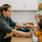 Two children sit at a table; one in a white shirt uses The Play Way's green tool to cut strawberries on a wooden board, focusing intently. The other wears yellow, watching with interest as strawberry pieces sit nearby. Part of the Feeding and Mealtimes: Helping You Get Started Program.