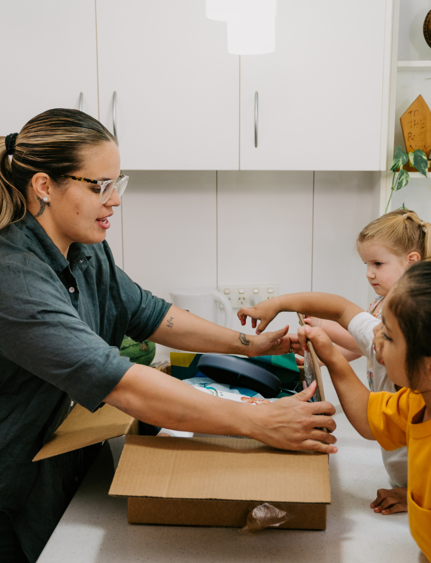Two children sit at a table; one in a white shirt uses The Play Way's green tool to cut strawberries on a wooden board, focusing intently. The other wears yellow, watching with interest as strawberry pieces sit nearby. Part of the Feeding and Mealtimes: Helping You Get Started Program.