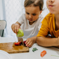 Two children sit at a table; one in a white shirt uses The Play Way's green tool to cut strawberries on a wooden board, focusing intently. The other wears yellow, watching with interest as strawberry pieces sit nearby. Part of the Feeding and Mealtimes: Helping You Get Started Program.