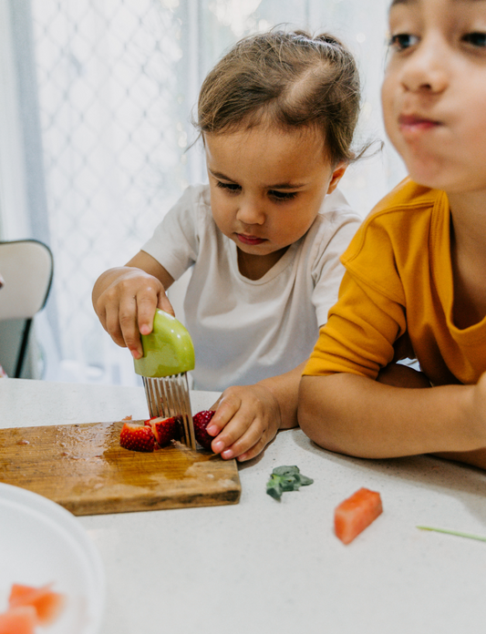 Two children sit at a table; one in a white shirt uses The Play Way's green tool to cut strawberries on a wooden board, focusing intently. The other wears yellow, watching with interest as strawberry pieces sit nearby. Part of the Feeding and Mealtimes: Helping You Get Started Program.