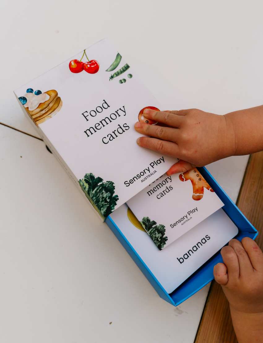 Two children sit at a table; one in a white shirt uses The Play Way's green tool to cut strawberries on a wooden board, focusing intently. The other wears yellow, watching with interest as strawberry pieces sit nearby. Part of the Feeding and Mealtimes: Helping You Get Started Program.