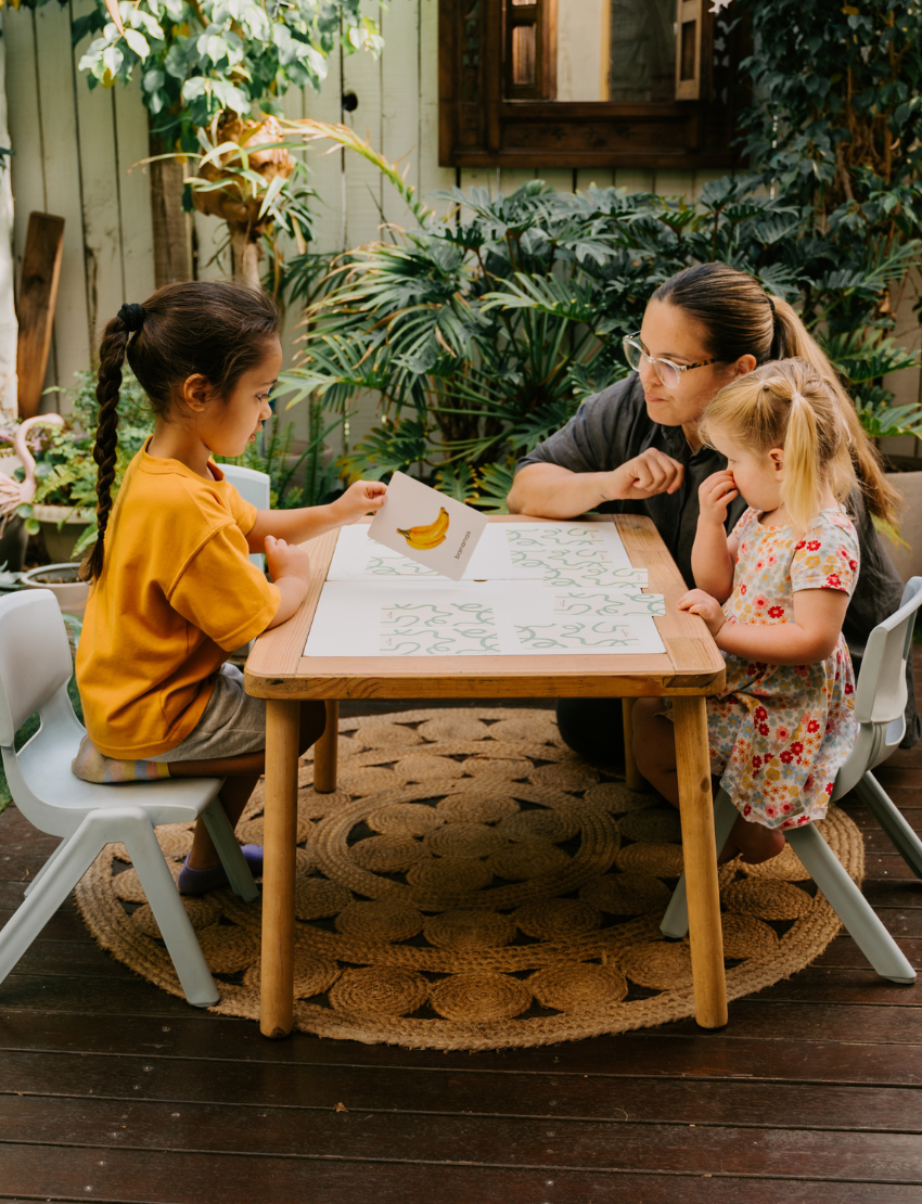Two children sit at a table; one in a white shirt uses The Play Way's green tool to cut strawberries on a wooden board, focusing intently. The other wears yellow, watching with interest as strawberry pieces sit nearby. Part of the Feeding and Mealtimes: Helping You Get Started Program.