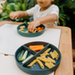 Two children sit at a table; one in a white shirt uses The Play Way's green tool to cut strawberries on a wooden board, focusing intently. The other wears yellow, watching with interest as strawberry pieces sit nearby. Part of the Feeding and Mealtimes: Helping You Get Started Program.