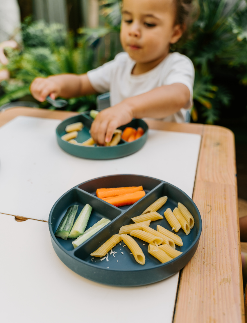 Two children sit at a table; one in a white shirt uses The Play Way's green tool to cut strawberries on a wooden board, focusing intently. The other wears yellow, watching with interest as strawberry pieces sit nearby. Part of the Feeding and Mealtimes: Helping You Get Started Program.