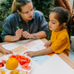 Two children sit at a table; one in a white shirt uses The Play Way's green tool to cut strawberries on a wooden board, focusing intently. The other wears yellow, watching with interest as strawberry pieces sit nearby. Part of the Feeding and Mealtimes: Helping You Get Started Program.