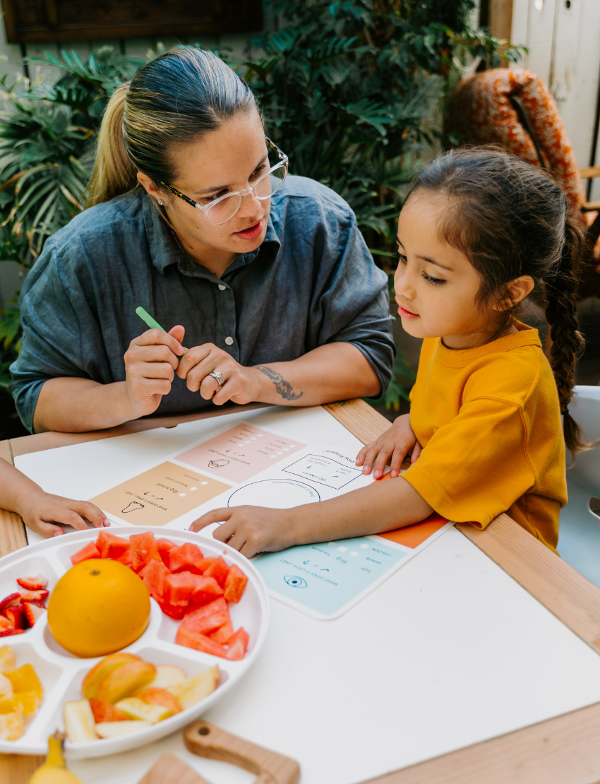 Two children sit at a table; one in a white shirt uses The Play Way's green tool to cut strawberries on a wooden board, focusing intently. The other wears yellow, watching with interest as strawberry pieces sit nearby. Part of the Feeding and Mealtimes: Helping You Get Started Program.