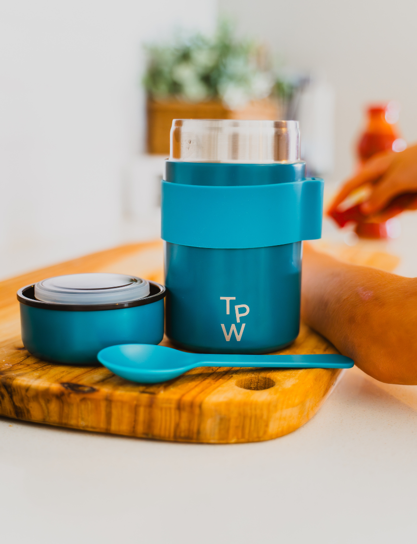 A young girl with long hair smiles while sitting at a wooden table on a terrace, holding The Play Way's Lunch Box Program—metal with multiple compartments. Two small round containers with blue lids are also on the table.