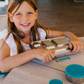 A young girl with long hair smiles while sitting at a wooden table on a terrace, holding The Play Way's Lunch Box Program—metal with multiple compartments. Two small round containers with blue lids are also on the table.