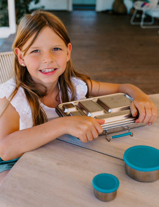 A young girl with long hair smiles while sitting at a wooden table on a terrace, holding The Play Way's Lunch Box Program—metal with multiple compartments. Two small round containers with blue lids are also on the table.