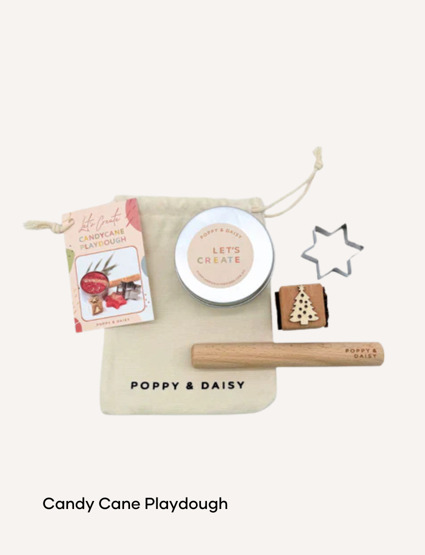 A hand holds a small tin of Lavender Lip Balm. Below it, a cloth pouch labeled "Poppy & Daisy" contains a glass vial, a small wooden stick, and a card with floral illustrations. The background reveals a knitted blanket.