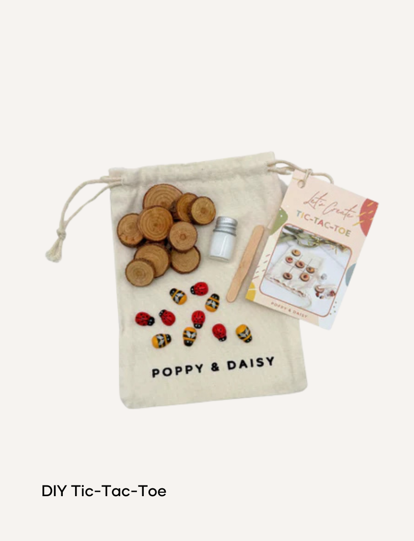 A hand holds a small tin of Lavender Lip Balm. Below it, a cloth pouch labeled "Poppy & Daisy" contains a glass vial, a small wooden stick, and a card with floral illustrations. The background reveals a knitted blanket.