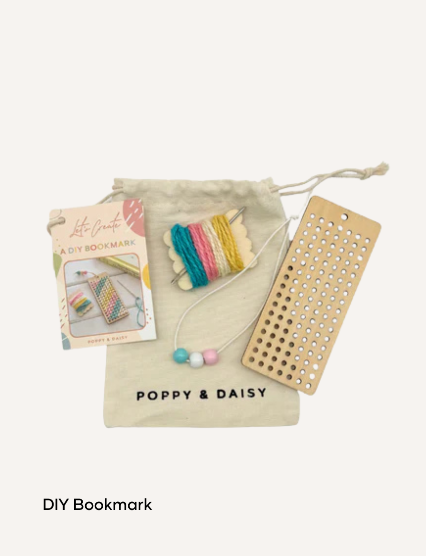 A hand holds a small tin of Lavender Lip Balm. Below it, a cloth pouch labeled "Poppy & Daisy" contains a glass vial, a small wooden stick, and a card with floral illustrations. The background reveals a knitted blanket.