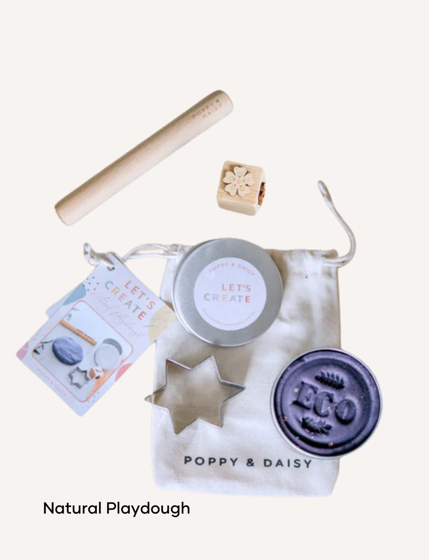 A hand holds a small tin of Lavender Lip Balm. Below it, a cloth pouch labeled "Poppy & Daisy" contains a glass vial, a small wooden stick, and a card with floral illustrations. The background reveals a knitted blanket.